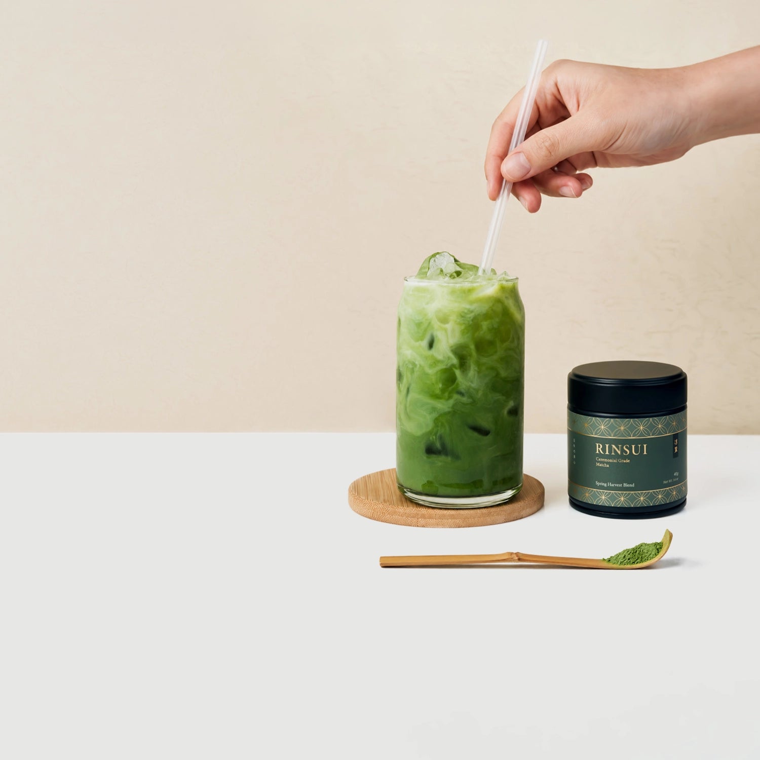 A hand holds a clear straw above an iced green drink in a glass on a wooden coaster, with a dark green RINSUI jar and a small bamboo spoon of green powder on a white surface against a beige background.