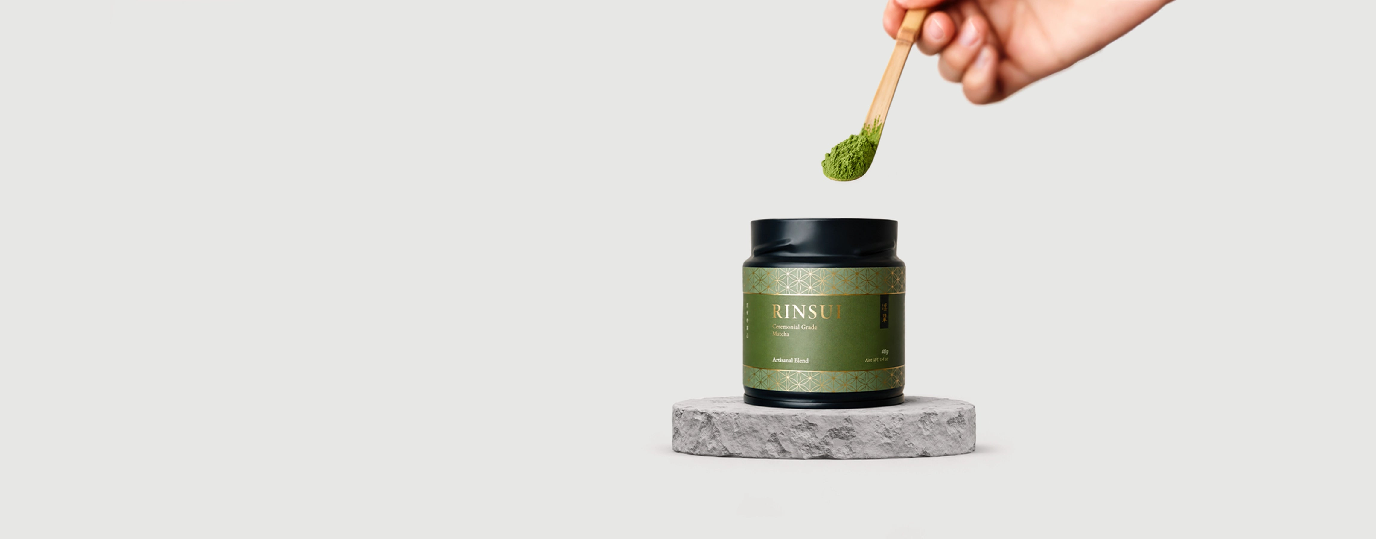 A hand holds a wooden spoon above a green cosmetic jar with a black lid, set on a gray marble pedestal against a light gray background. RINSUI ceremonial grade matcha artisanal blend 